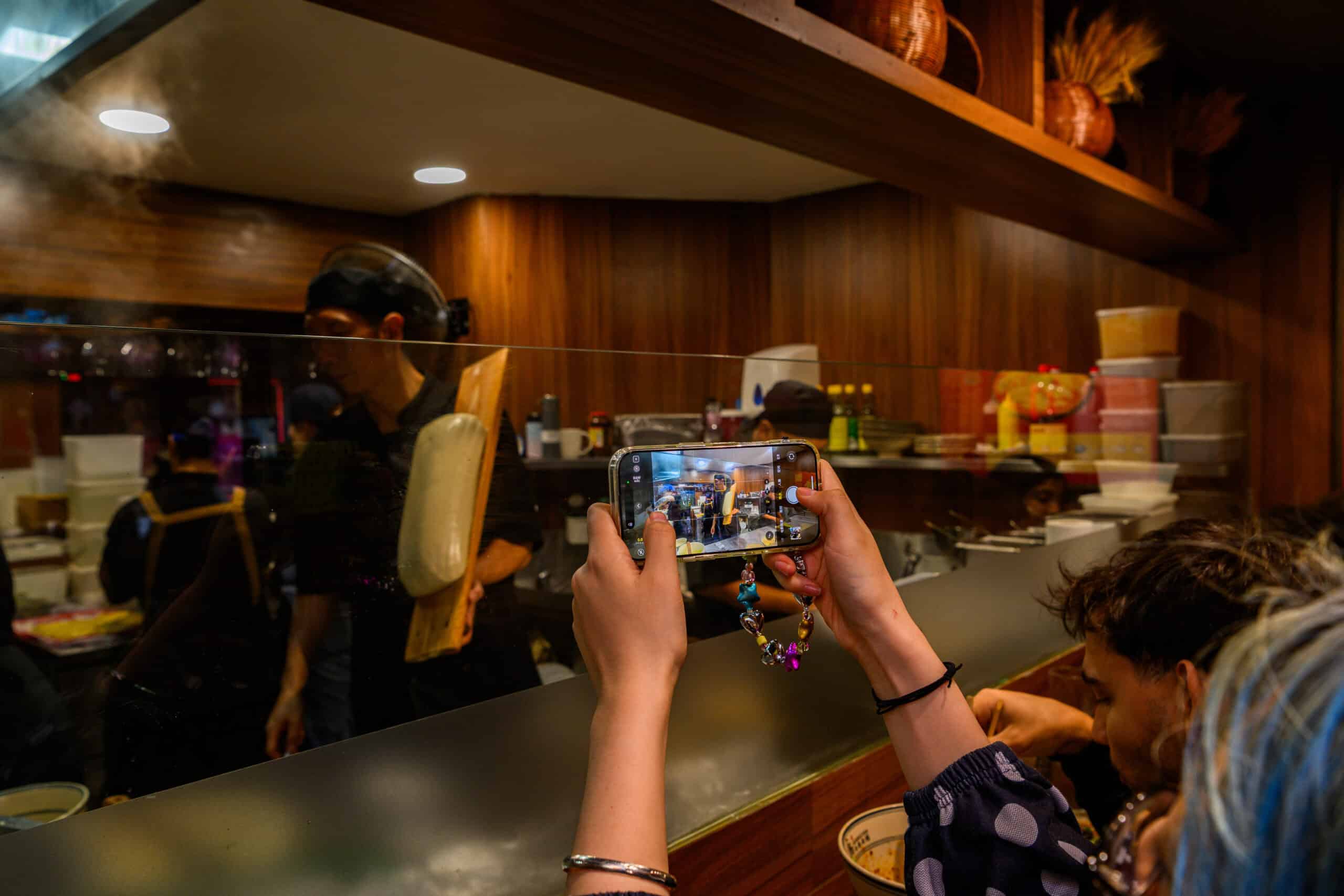 Customer taking a photo inside Noodle Inn restaurant with staff preparing food in the background.
