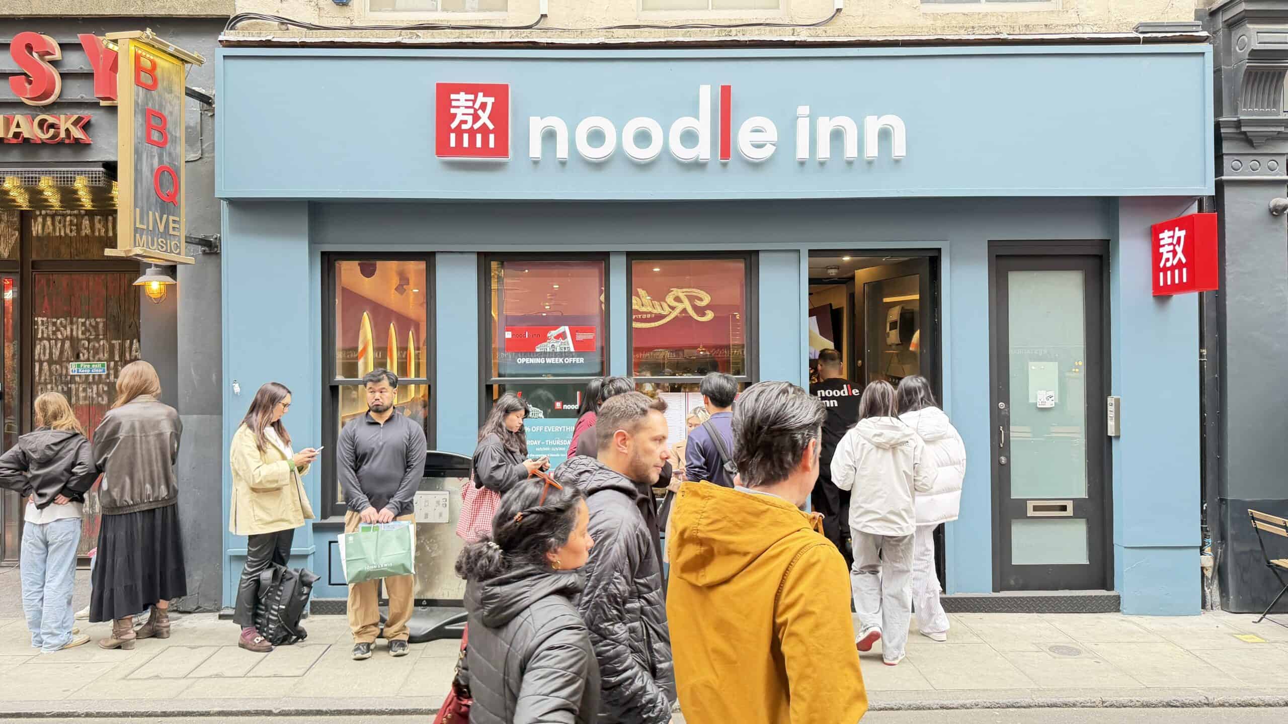 Vibrant Noodle Inn restaurant exterior with customers waiting outside on a busy street.